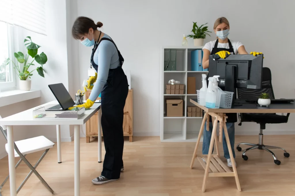 Two cleaning ladies in a room; one cleans a desk while the other cleans a monitor on a separate desk.